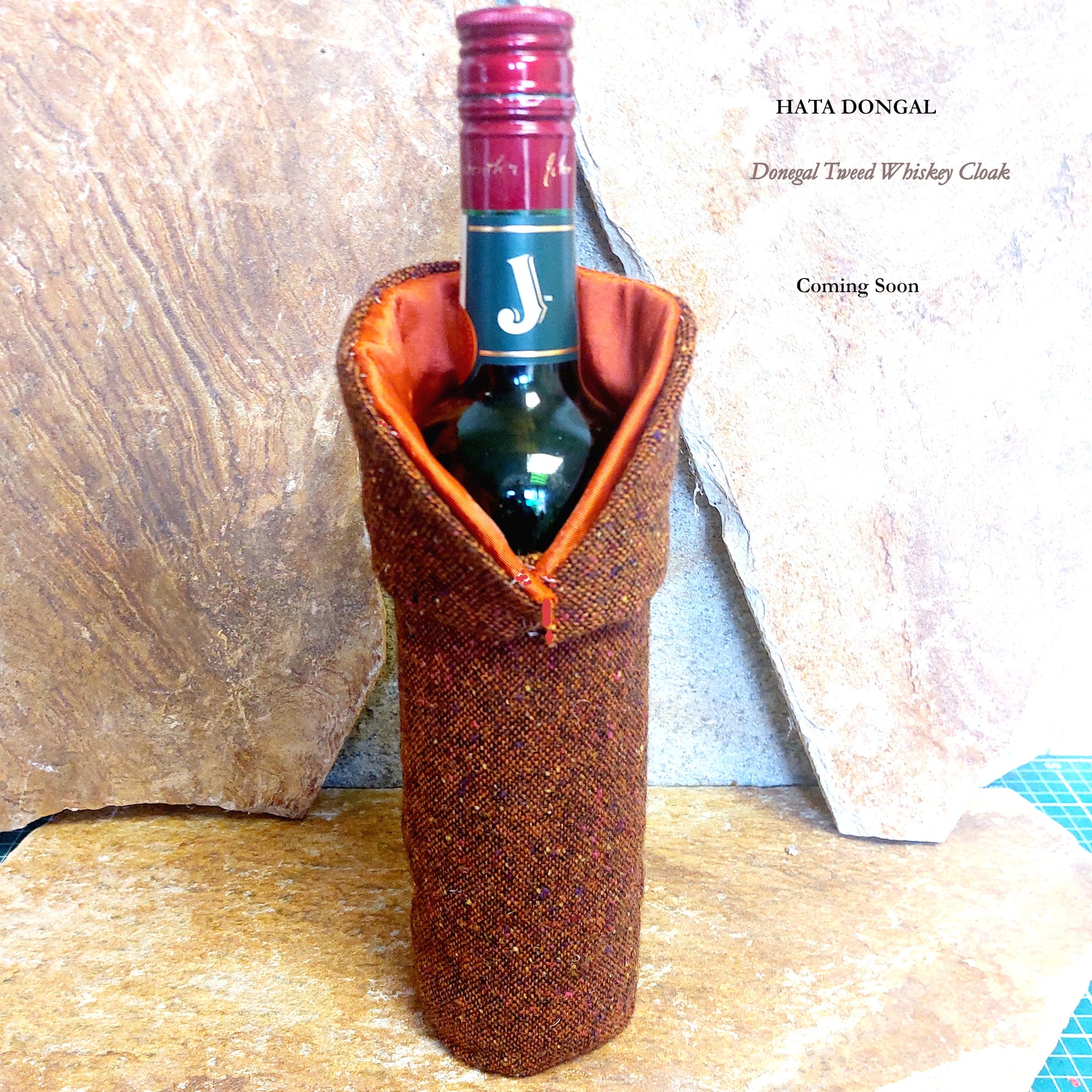 Donegal Tweed Whiskey Cloak by Hata Donegal, handcrafted in County Donegal from authentic Donegal tweed with contrasting orange lining, shown on a Jameson whiskey bottle against Donegal quartzite stone.
